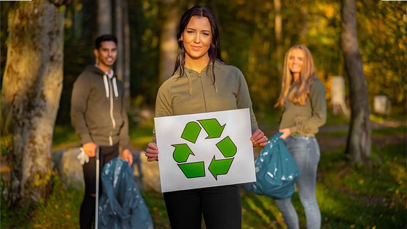 Girl with a recycling sign with two people behind her picking up trash.