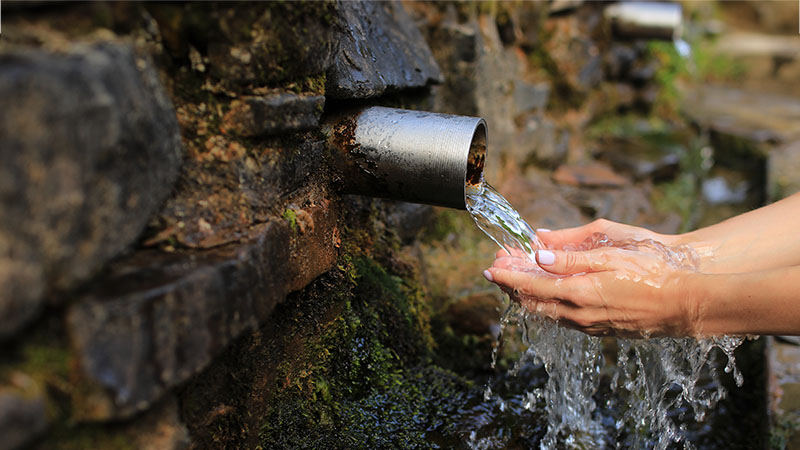 Water flowing out of a pipe in the side of rocks in to a person's hands.